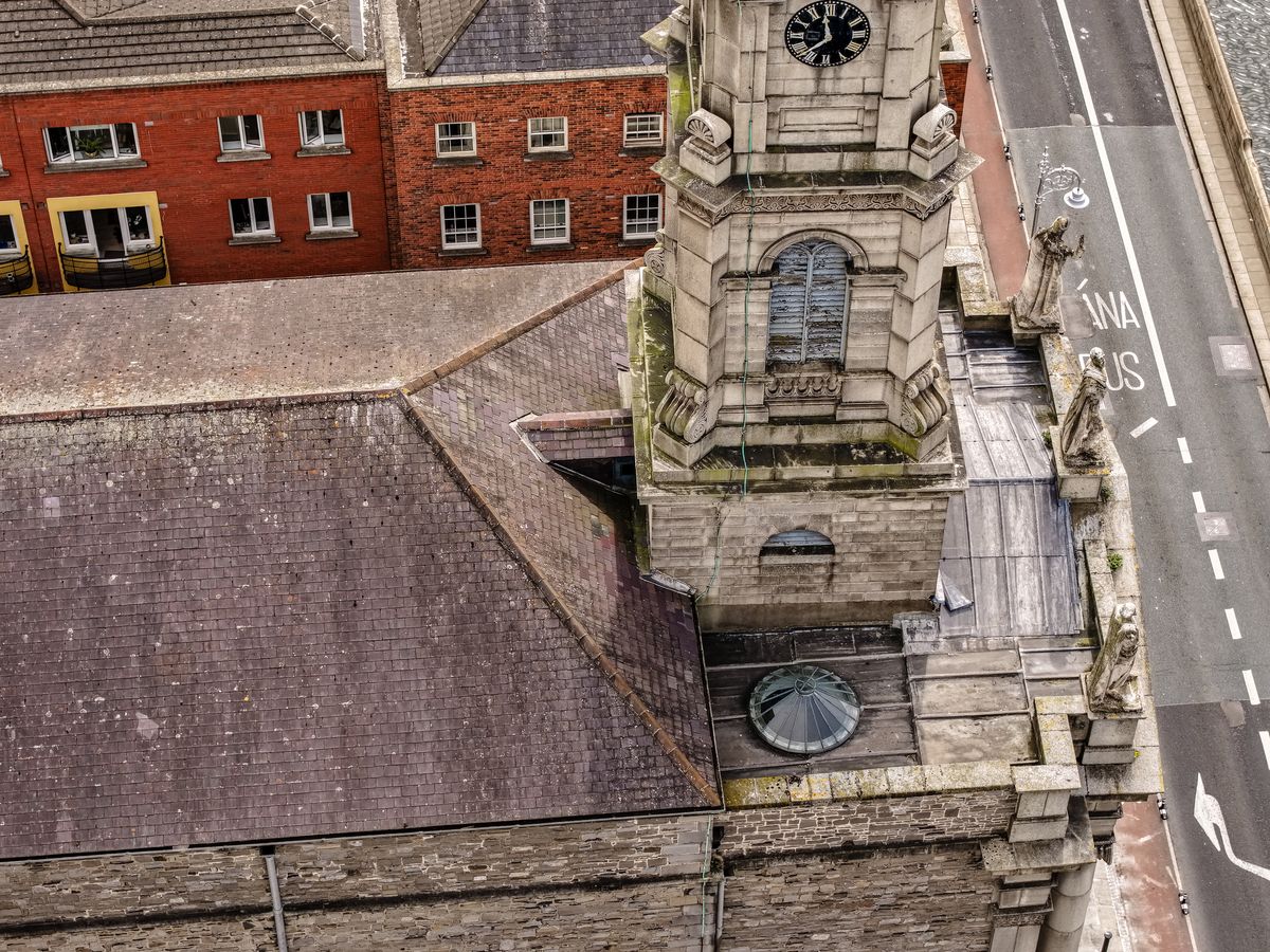 Aerial view of an ornate stone clock-tower facade beside a slate roof and red-brick building, surveyed before cleaning