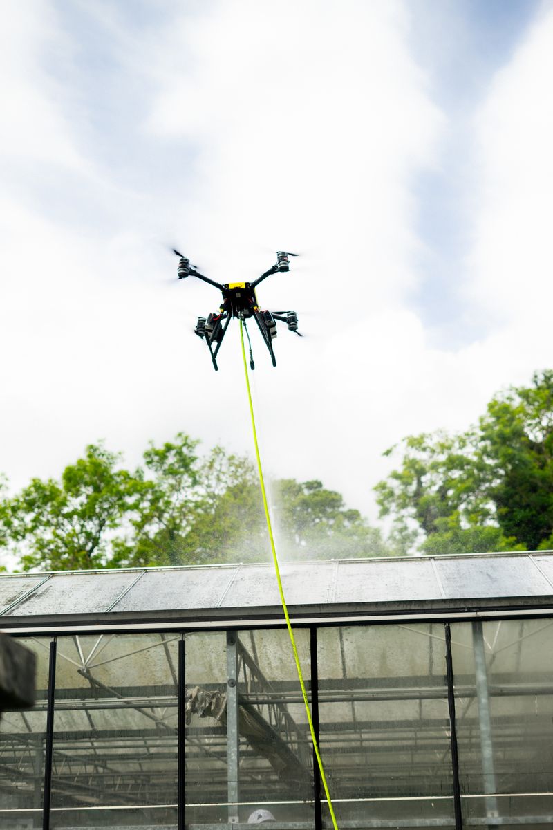 Tethered drone spraying water to clean a glasshouse roof during a roof and facade wash