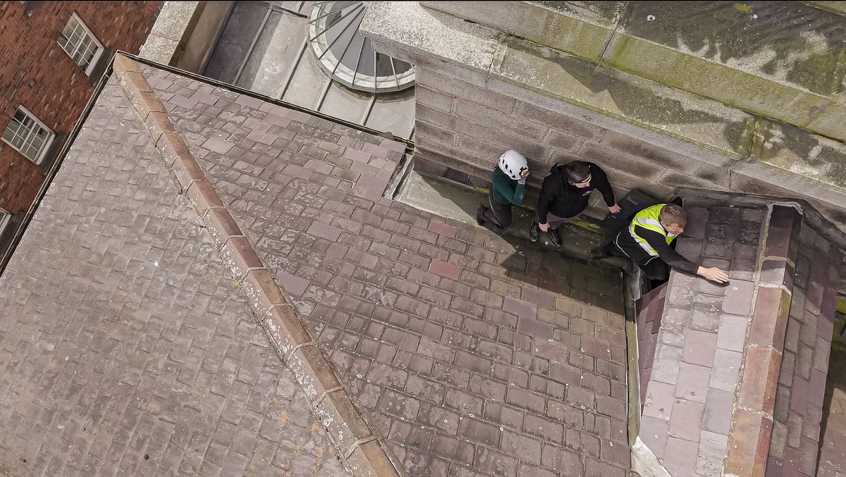 Aerial view looking down on three inspectors carrying out a condition survey on a pitched slate roof beside a stone tower
